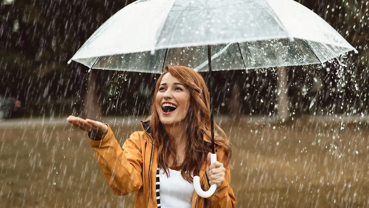 Waist up portrait of delighted girl standing outside and hiding under umbrella. She is stretching hand catching water drops with joy