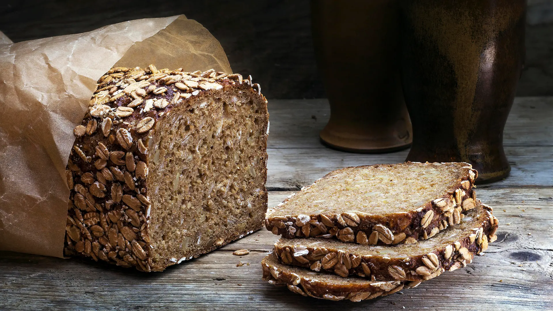 wholegrain rye bread with seeds on a weathered wooden board, rustic earthenware in the dark background