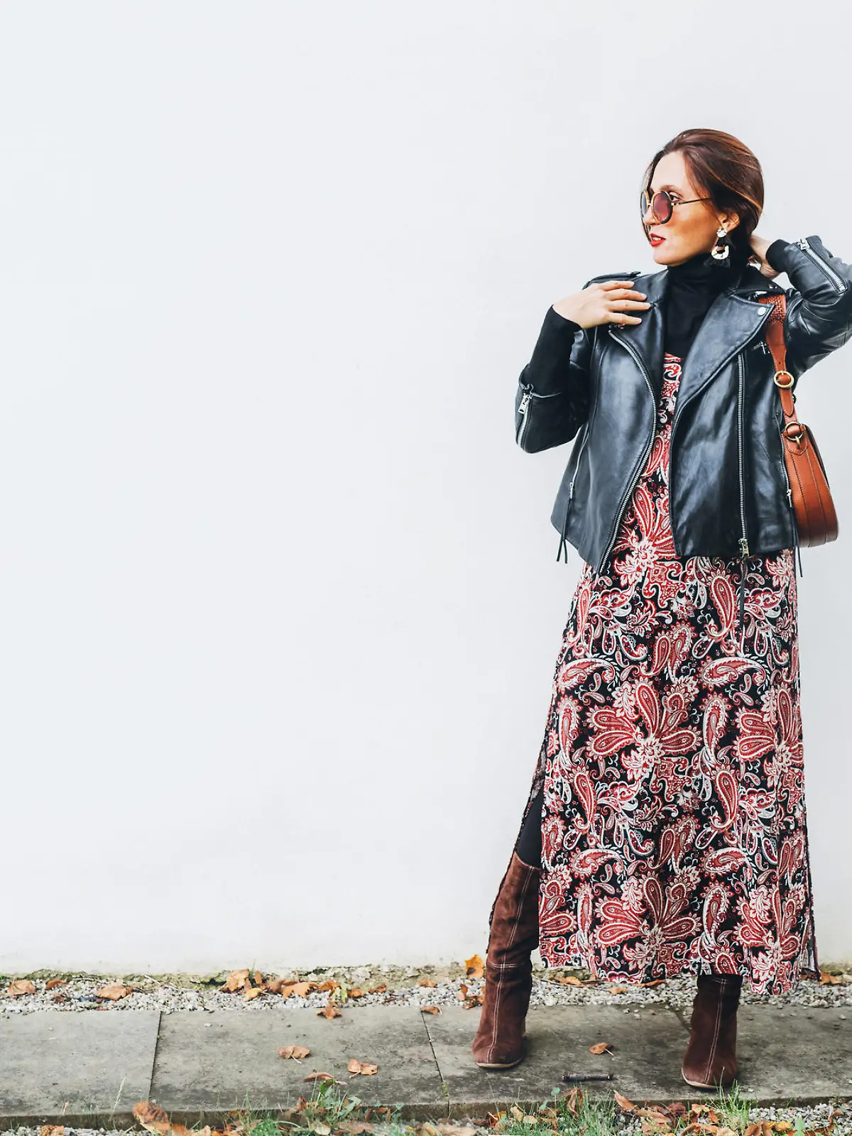 Smiling female dressed boho fashion style colorful long dress with black leather biker jacket with brown leather flap bag posing on the white wall background.