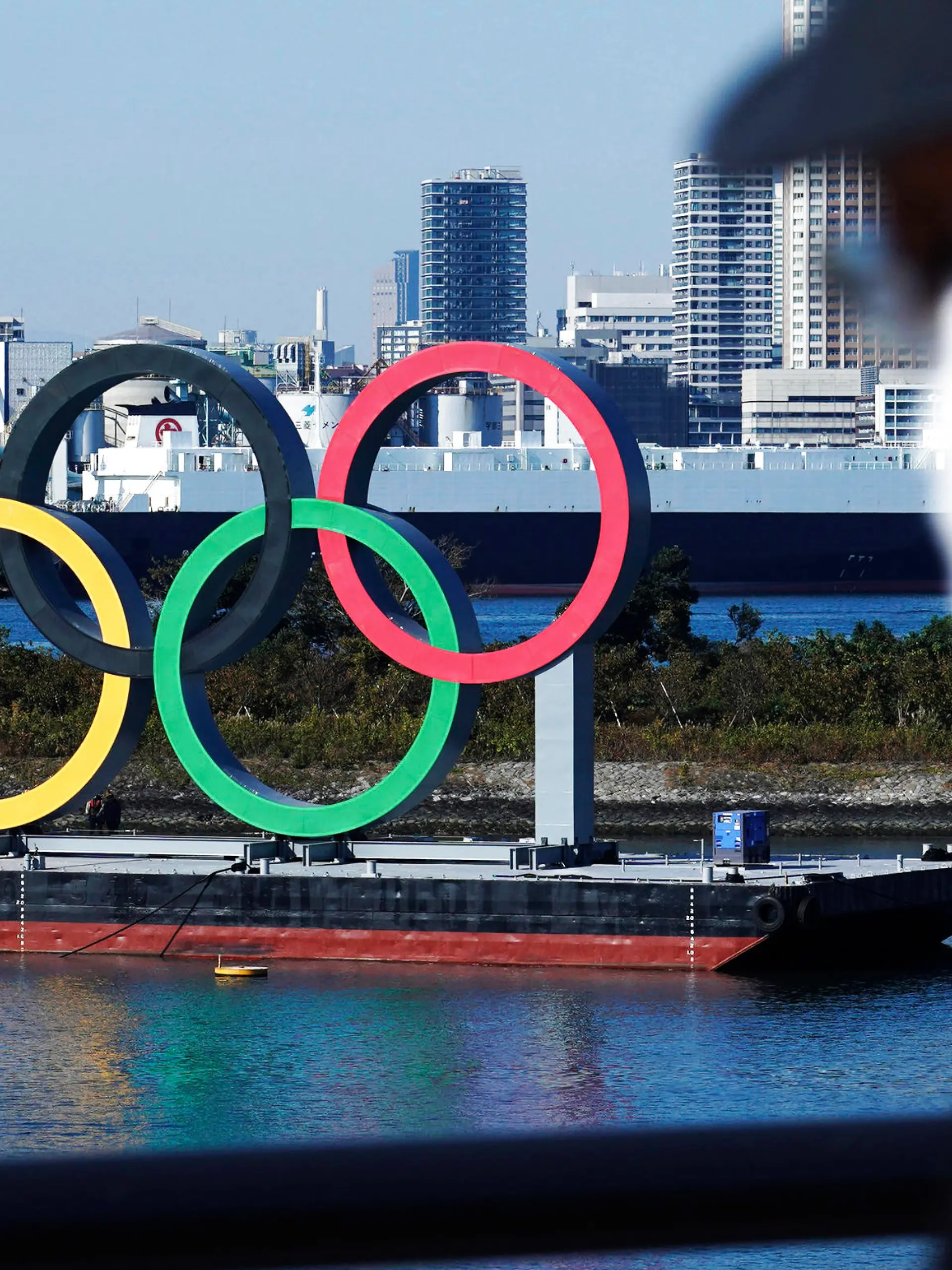 ARCHIV - 01.12.2020, Japan, Tokio: Ein Mann, der eine schützende Gesichtsmaske trägt, um die Ausbreitung des Coronavirus einzudämmen, geht nahe der Olympischen Ringe in der Bucht von Tokio, nachdem diese vor vier Monaten zur Wartung entfernt wurden, kurz nachdem die Olympischen Spiele in Tokio wegen der Corona-Pandemie auf nächstes Jahr verschoben worden waren. Die Olympischen Spiele sollen vom 23. Juli bis zum 8. August 2021 in Tokio stattfinden. (zu dpa: Zu 100 Tage vor Olympia am 14. April) Foto: Eugene Hoshiko/AP/dpa +++ dpa-Bildfunk +++
