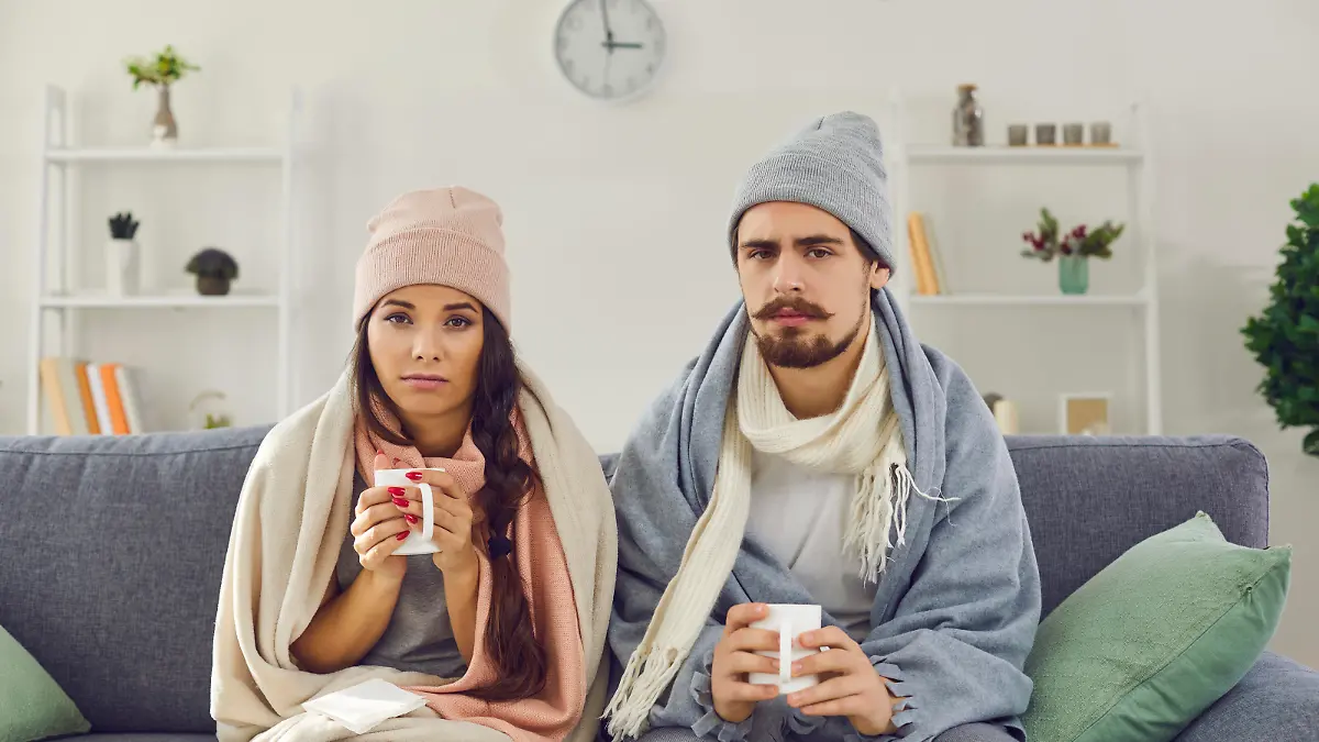 Dissatisfied young couple having problem with central heating, sitting on sofa at home, freezing, drinking hot tea trying to warm up. Sick man and woman wrapped in blankets suffering from cold or flu