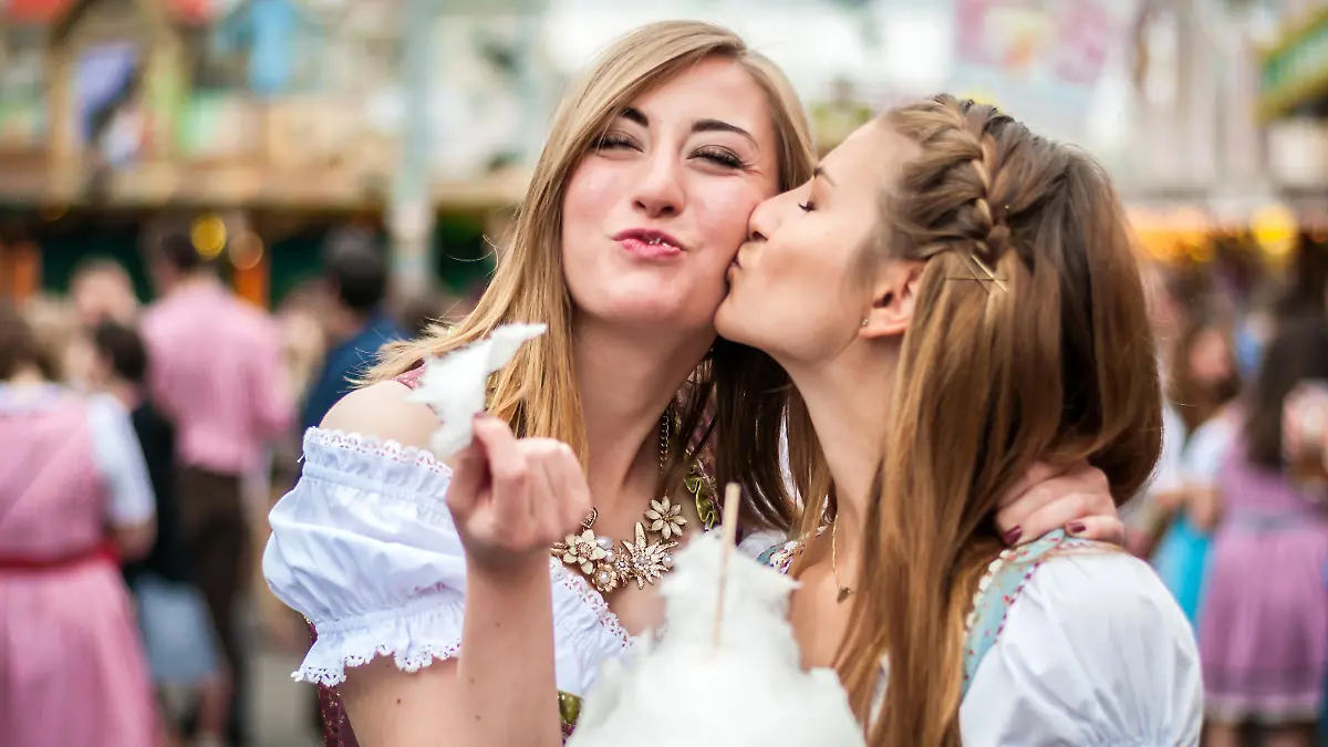 Two young women in traditional Bavarian clothes, dirndl or tracht, kissing with cotton candy floss at the Oktoberfest