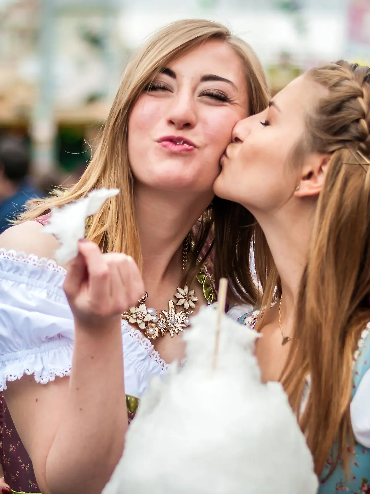 Two young women in traditional Bavarian clothes, dirndl or tracht, kissing with cotton candy floss at the Oktoberfest