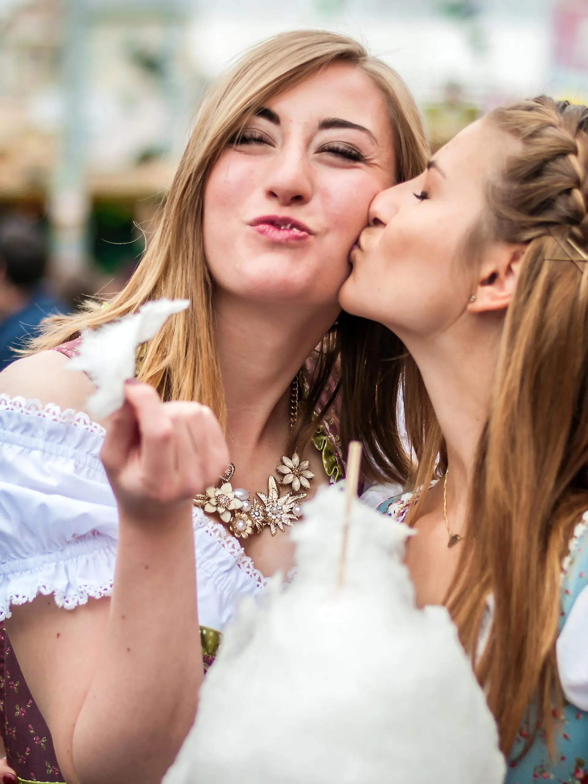 Two young women in traditional Bavarian clothes, dirndl or tracht, kissing with cotton candy floss at the Oktoberfest