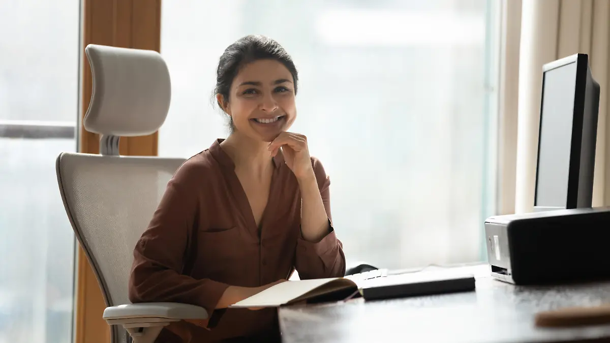 Smiling attractive Indian businesswoman sitting at desk in modern home office pose for camera feel confident, satisfied by business growth. Portrait of female freelancer, motivated student or employee