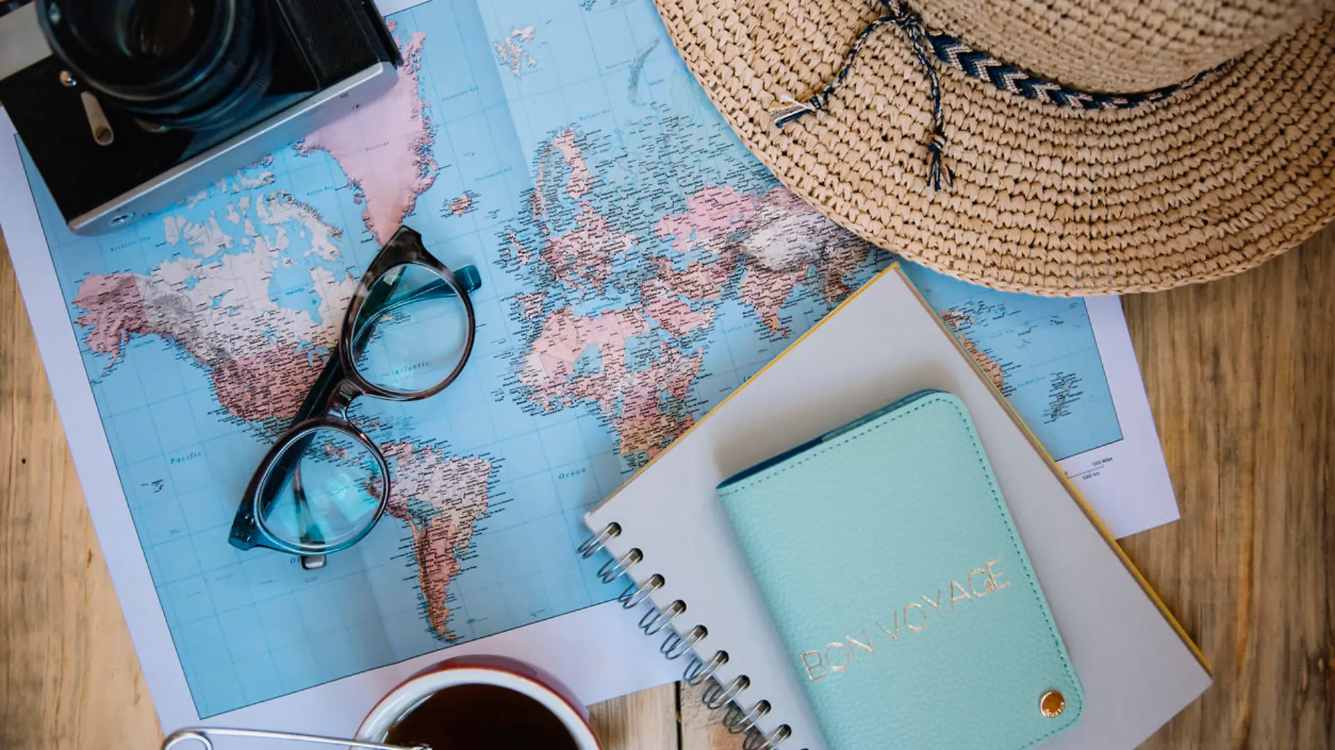 Travel preparations essentials. World map, cup of tea, vintage film camera, hat, passport, notebook and stylish glasses on the old rustic wooden table background, flat lay