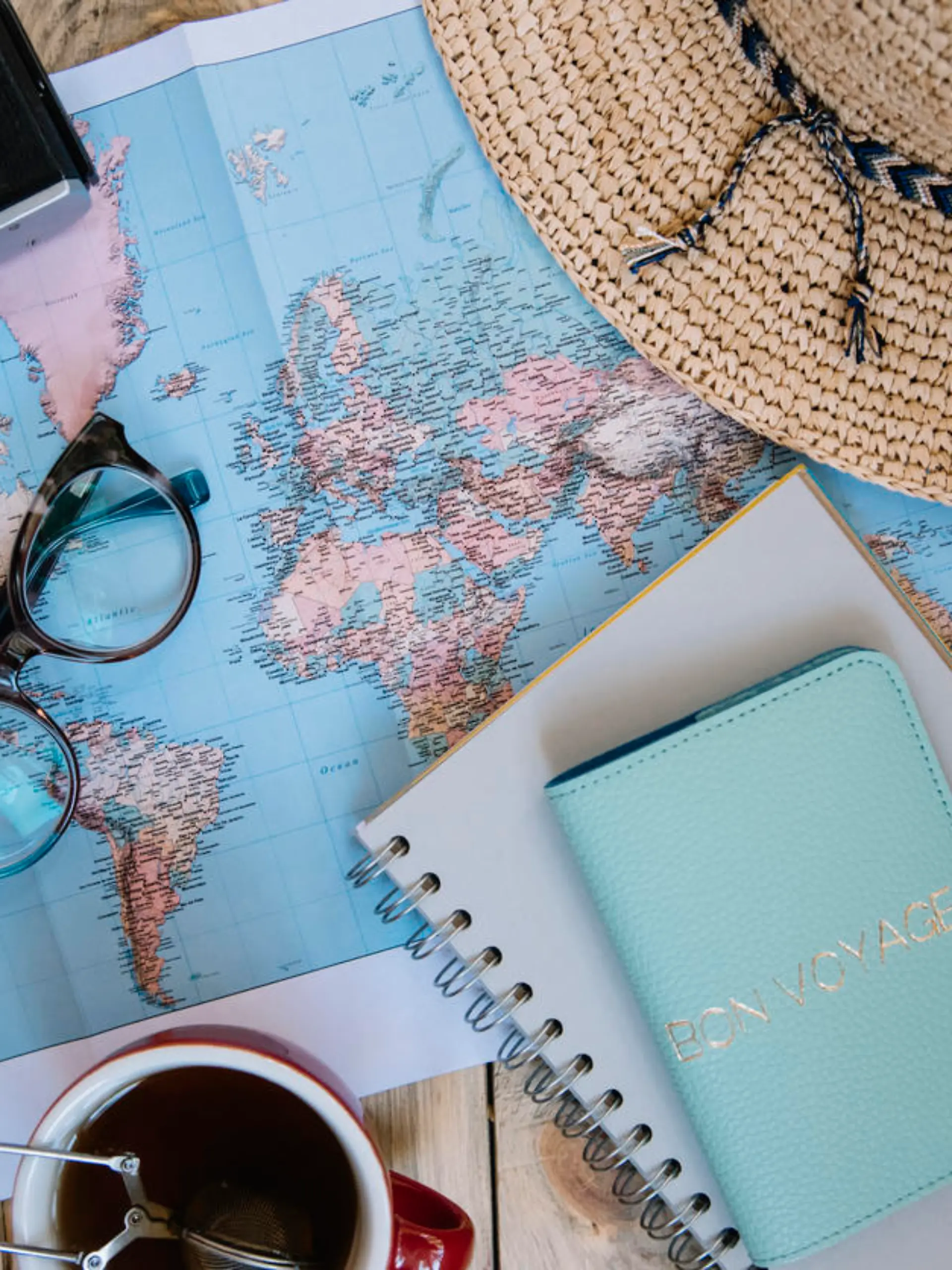 Travel preparations essentials. World map, cup of tea, vintage film camera, hat, passport, notebook and stylish glasses on the old rustic wooden table background, flat lay