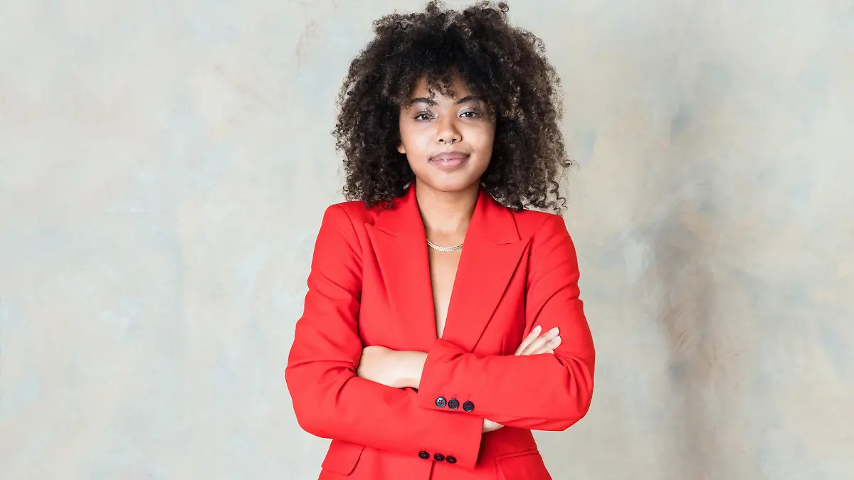 Fashion portrait of curly hair young woman wearing red suit, standing with arms crossed and smiling at camera. Studio shot on grey background.