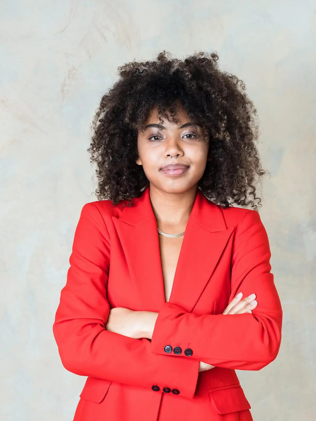 Fashion portrait of curly hair young woman wearing red suit, standing with arms crossed and smiling at camera. Studio shot on grey background.