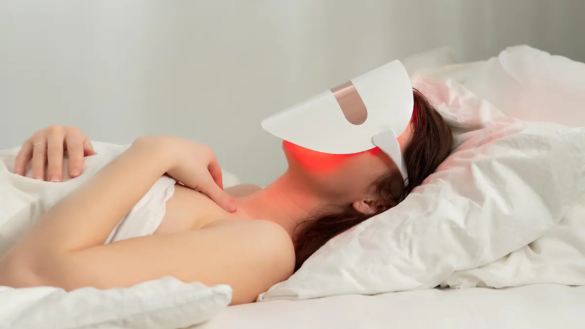 Woman lies in bed with led light therapy facial mask and relax. Home skincare and me time concept. Light rejuvenating mask for facial skin therapy. Photodynamic therapy mask on female face. Copy space