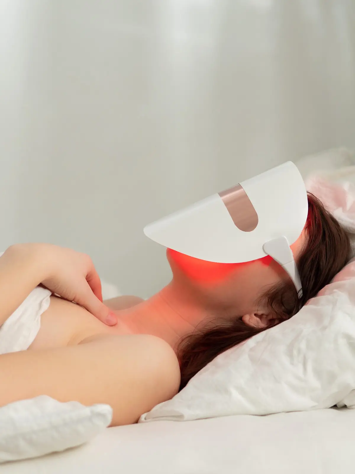 Woman lies in bed with led light therapy facial mask and relax. Home skincare and me time concept. Light rejuvenating mask for facial skin therapy. Photodynamic therapy mask on female face. Copy space