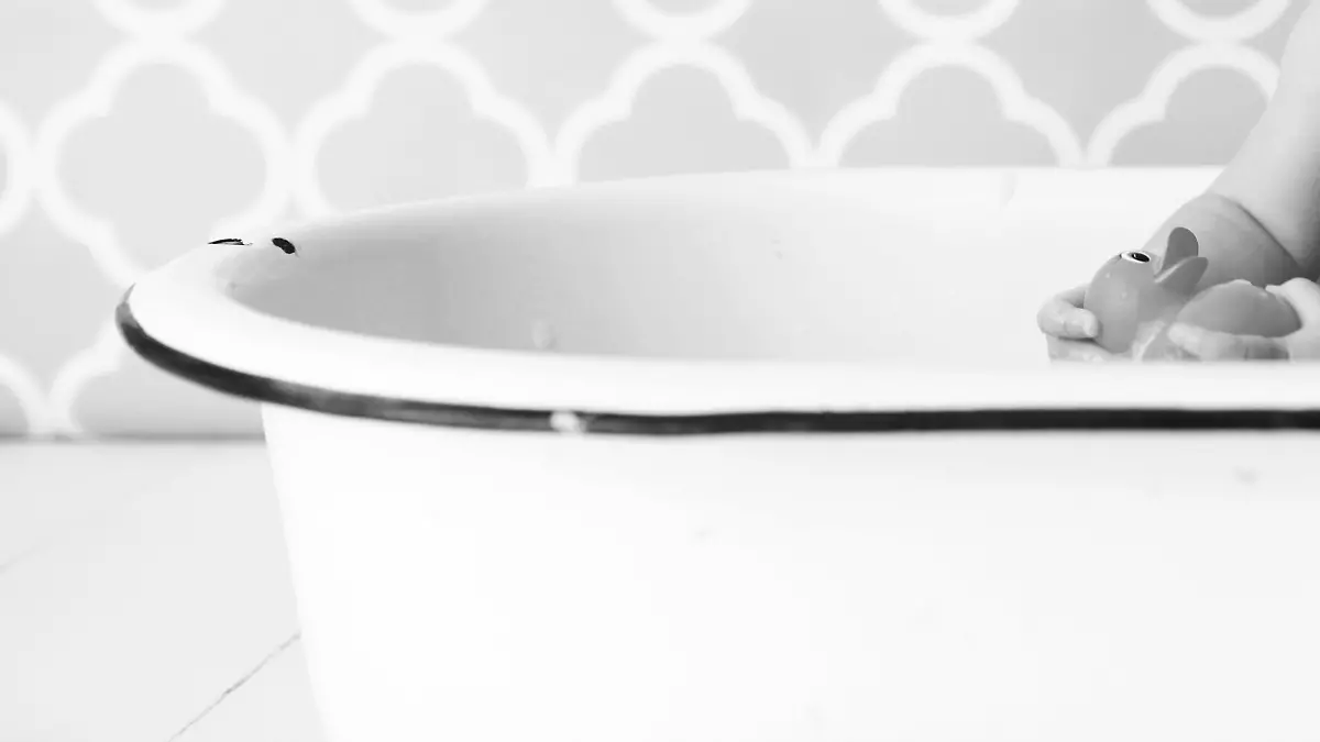 A baby looks at a rubber ducky as she sits in an antique bathtub. Copy space in vintage wallpaper.