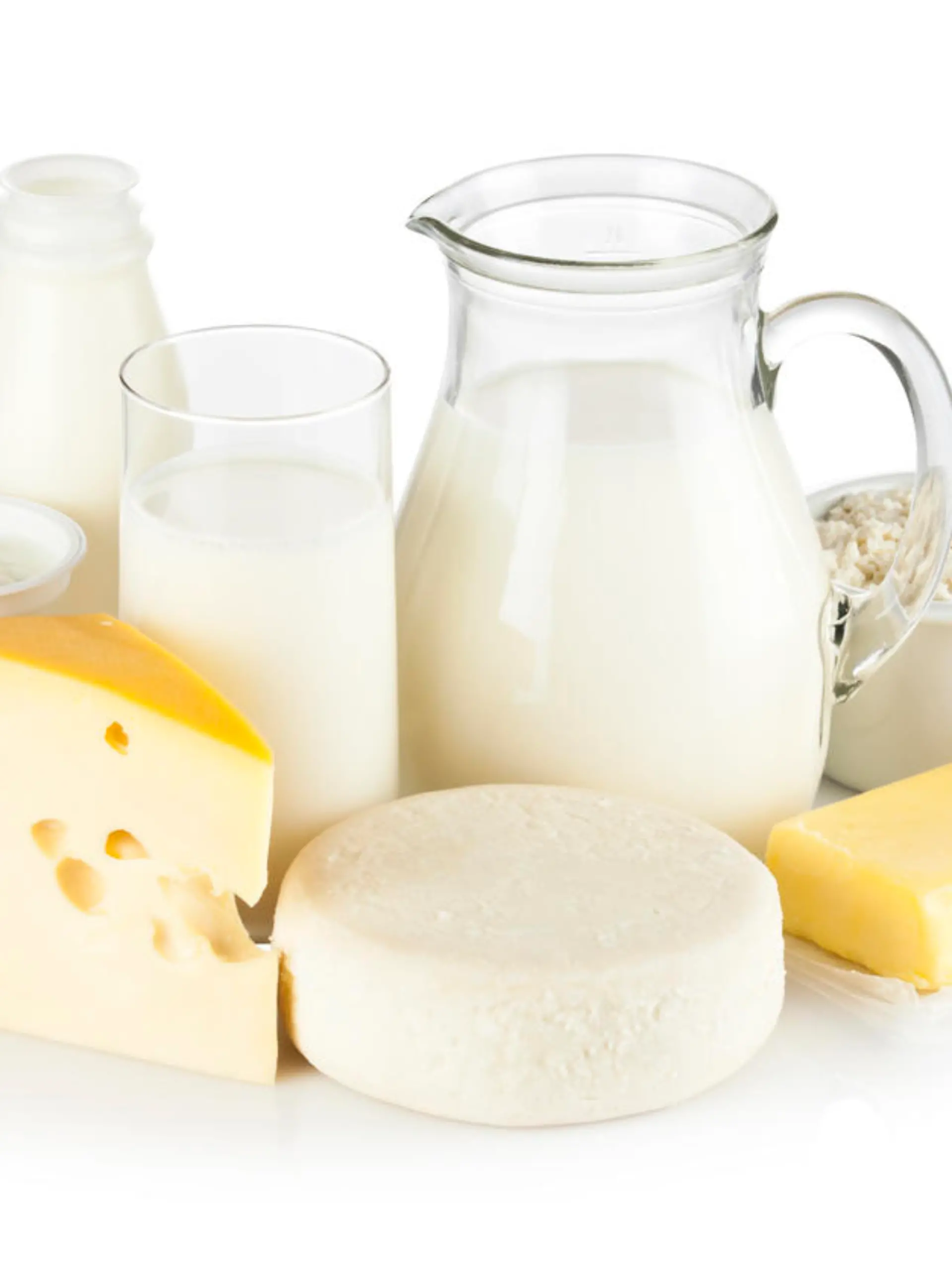 Image of dairy products shot on reflective white backdrop. Includes: milk, various types of cheese, butter, ricotta, milk cream and yogurt. A milk pitcher and a glass of milk are in the center while the rest of the products are around them. A soft shadow is visible in the foreground. The predominant color is white. DSRL studio shot with Canon EOS 5D Mk II