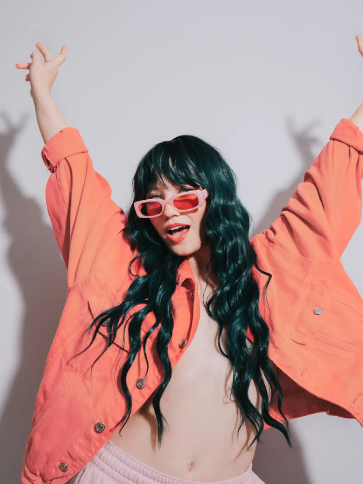 Cheeky emotional fashion young woman with long green color hair and trendy peach color lips, jacket and sunglasses dances on white background. Playful Hipster woman having fun. Color of the 2024 year