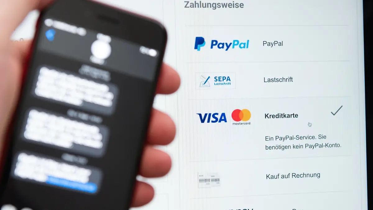 Ein Mann hält sein Smartphone zur Zwei-Faktoren-Authentifizierung in der Hand, während auf dem Bildschirm eines Laptops verschiedene Bezahlmethoden abgebildet sind.