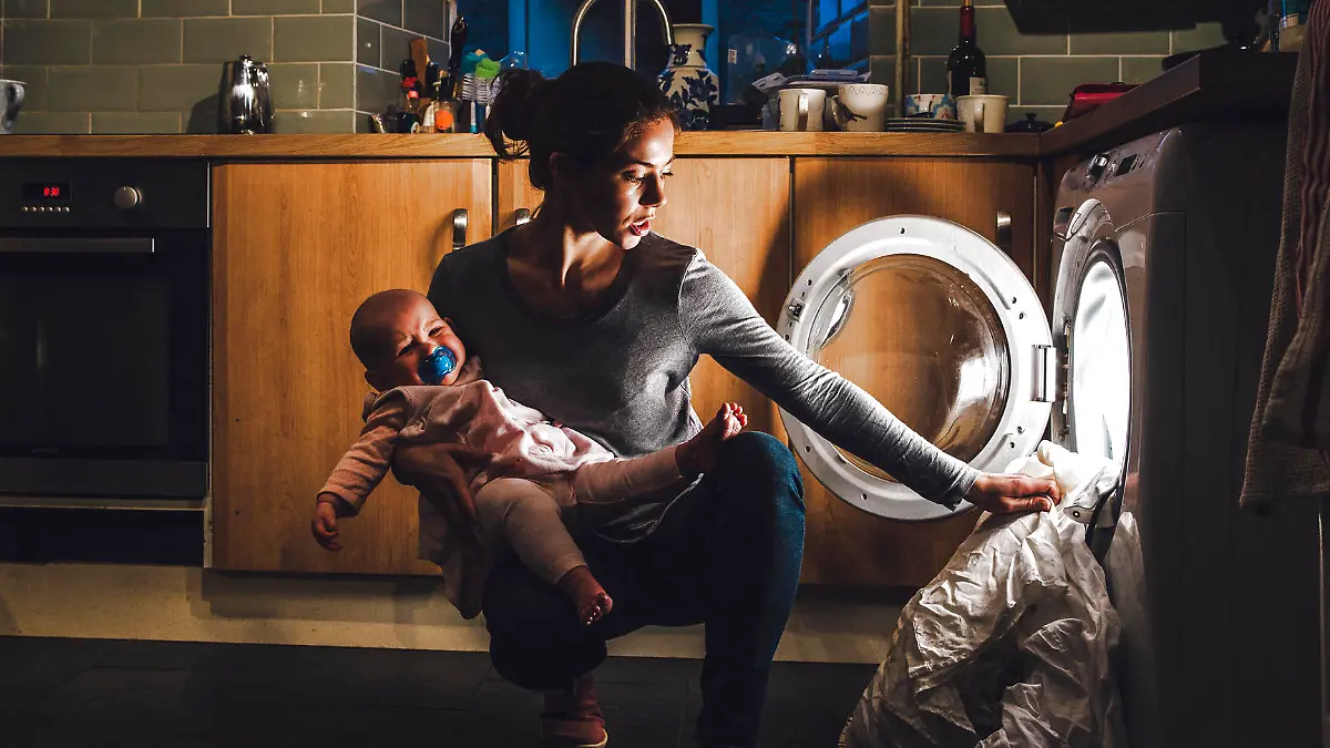 Young mother is holding her crying baby daughter on her hip while trying to load the washing machine.