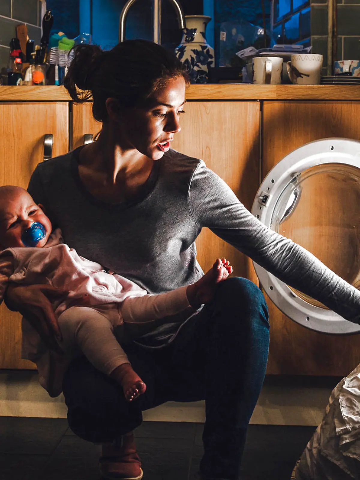 Young mother is holding her crying baby daughter on her hip while trying to load the washing machine.