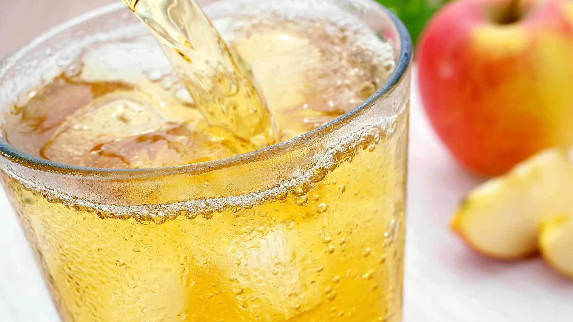 Ein Glas erfrischende eiskalte Apfelsaftschorle Apfelschorle mit Eisw?rfeln wird eingeschenkt. Das kalte Glas hat kondensierte Wassert?pfchen. Im Hintergrund ist ein Apfel und aufgeschnittene Apfelschnitze.