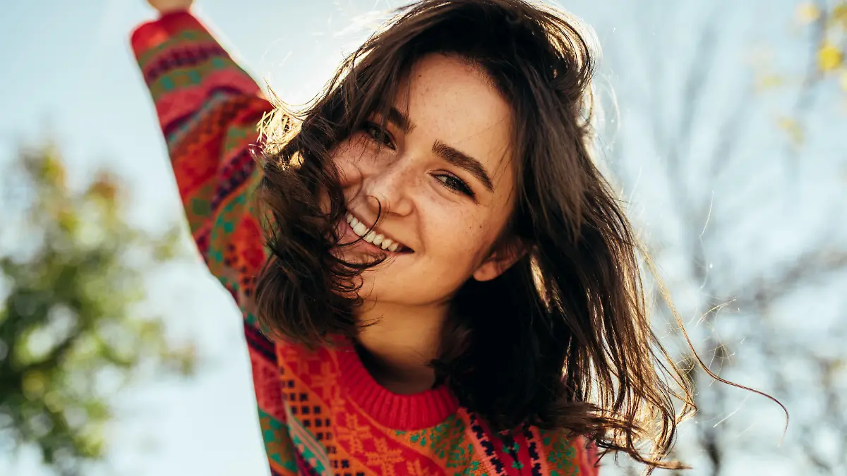 Bottom view close-up portrait of happy young woman smiling broadly with windy hair and freckles has joyful expression, wearing colorful knitted sweater with arms wide open, posing on nature sunlight