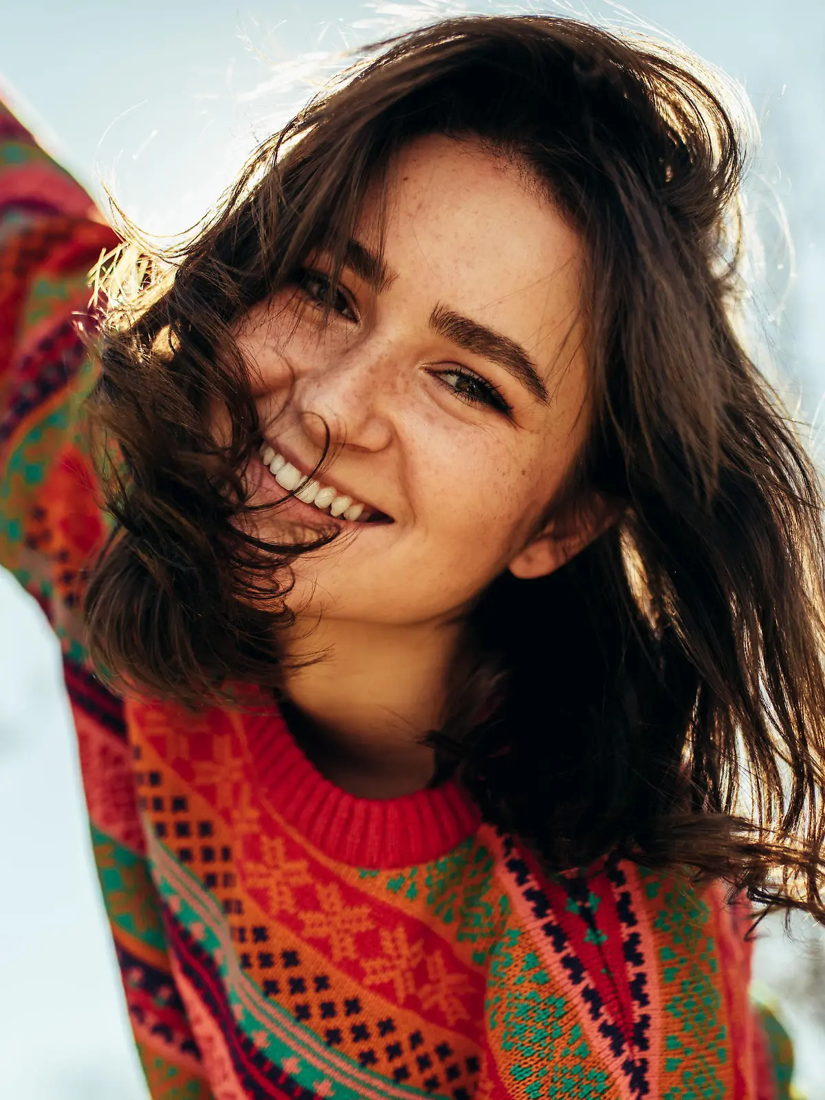 Bottom view close-up portrait of happy young woman smiling broadly with windy hair and freckles has joyful expression, wearing colorful knitted sweater with arms wide open, posing on nature sunlight