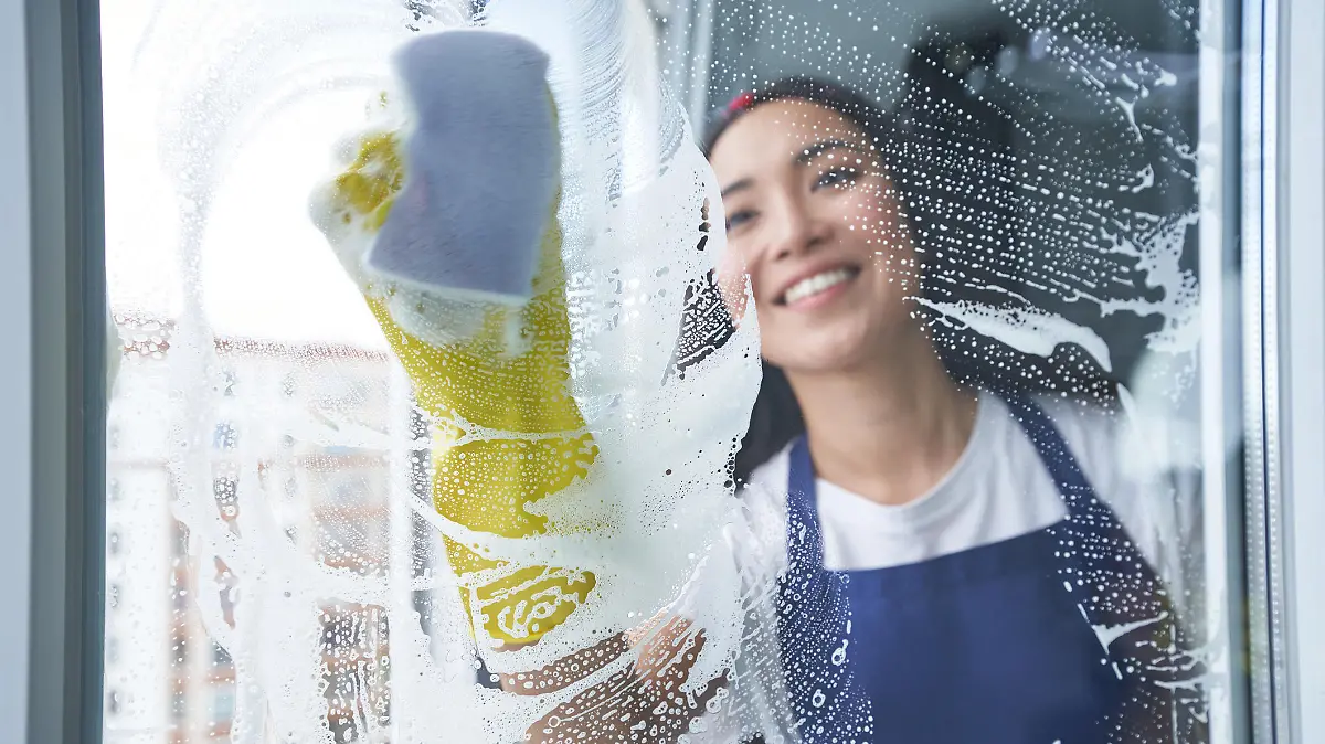 Cheerful young woman smiling while cleaning the window, glass surface using sponge. Housework and housekeeping, cleaning service concept. View through the glass