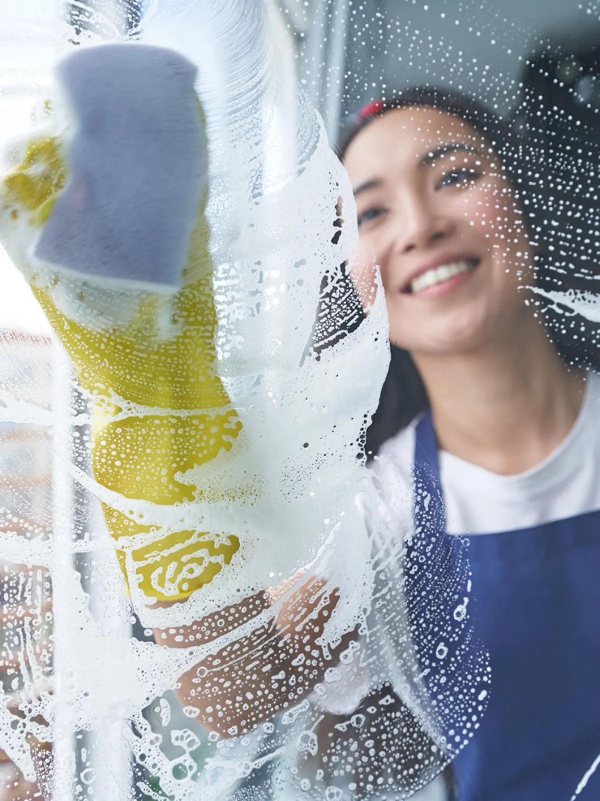 Cheerful young woman smiling while cleaning the window, glass surface using sponge. Housework and housekeeping, cleaning service concept. View through the glass