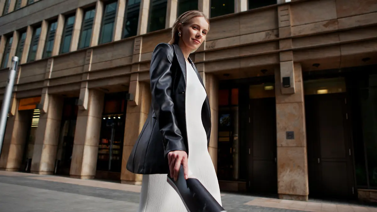 Portrait of blonde female fashion model in leather blazer jacket and white dress looking into the camera, walking in city. Young stylish woman outside.