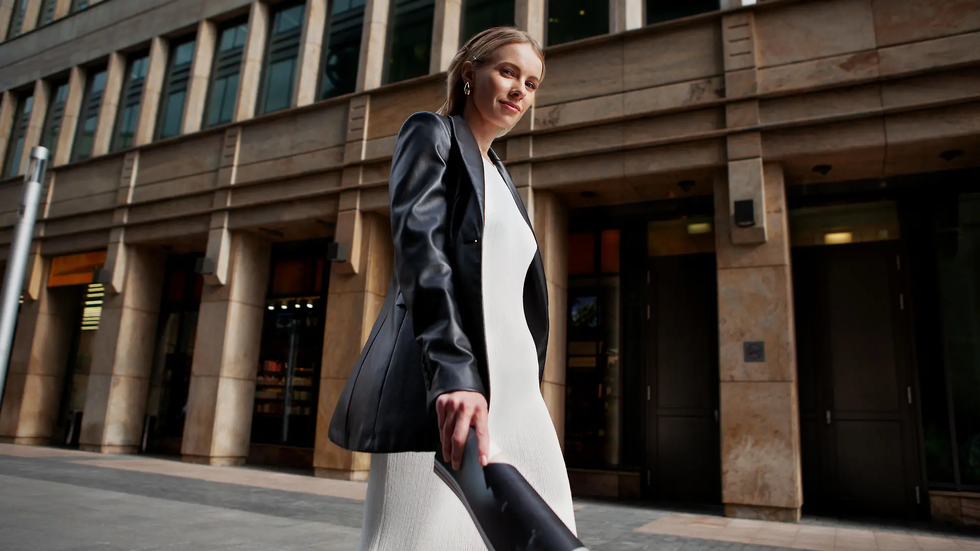 Portrait of blonde female fashion model in leather blazer jacket and white dress looking into the camera, walking in city. Young stylish woman outside.