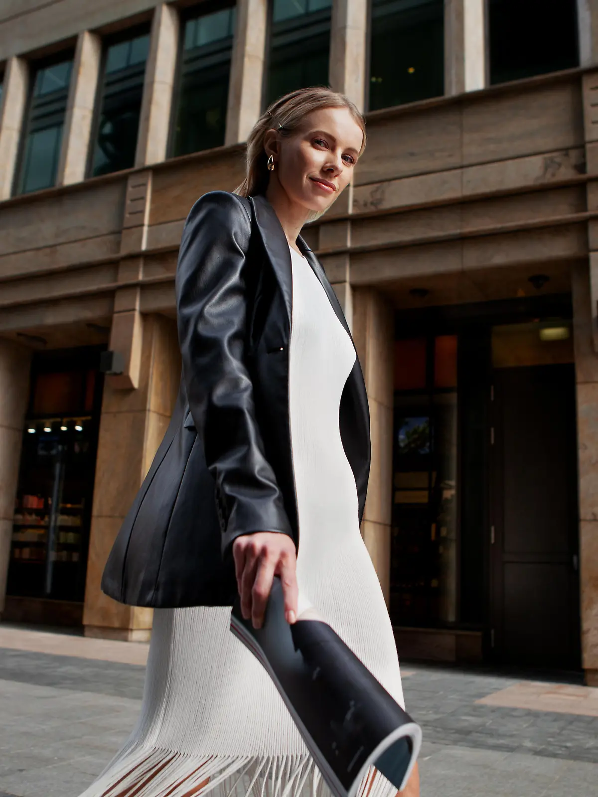 Portrait of blonde female fashion model in leather blazer jacket and white dress looking into the camera, walking in city. Young stylish woman outside.