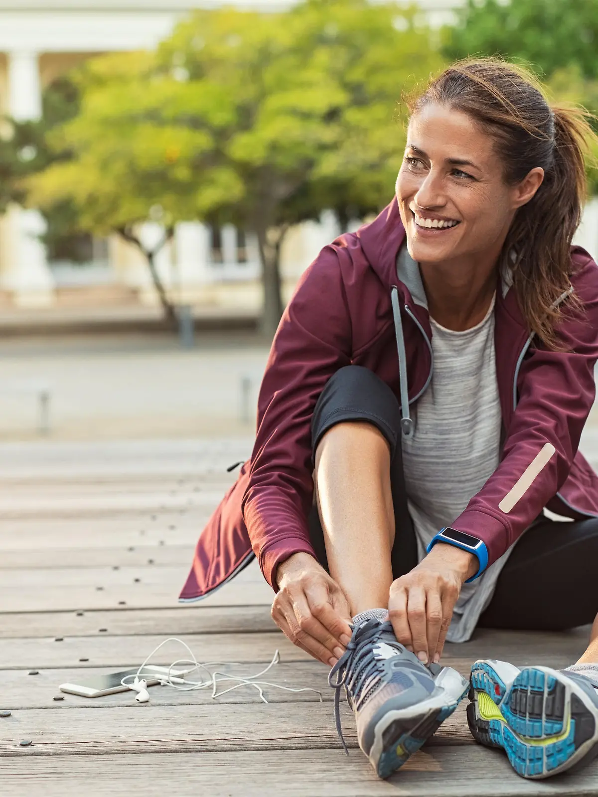 Mature fitness woman tie shoelaces on road. Cheerful runner sitting on floor on city streets with mobile and earphones wearing sport shoes. Active latin woman tying shoe lace before running.