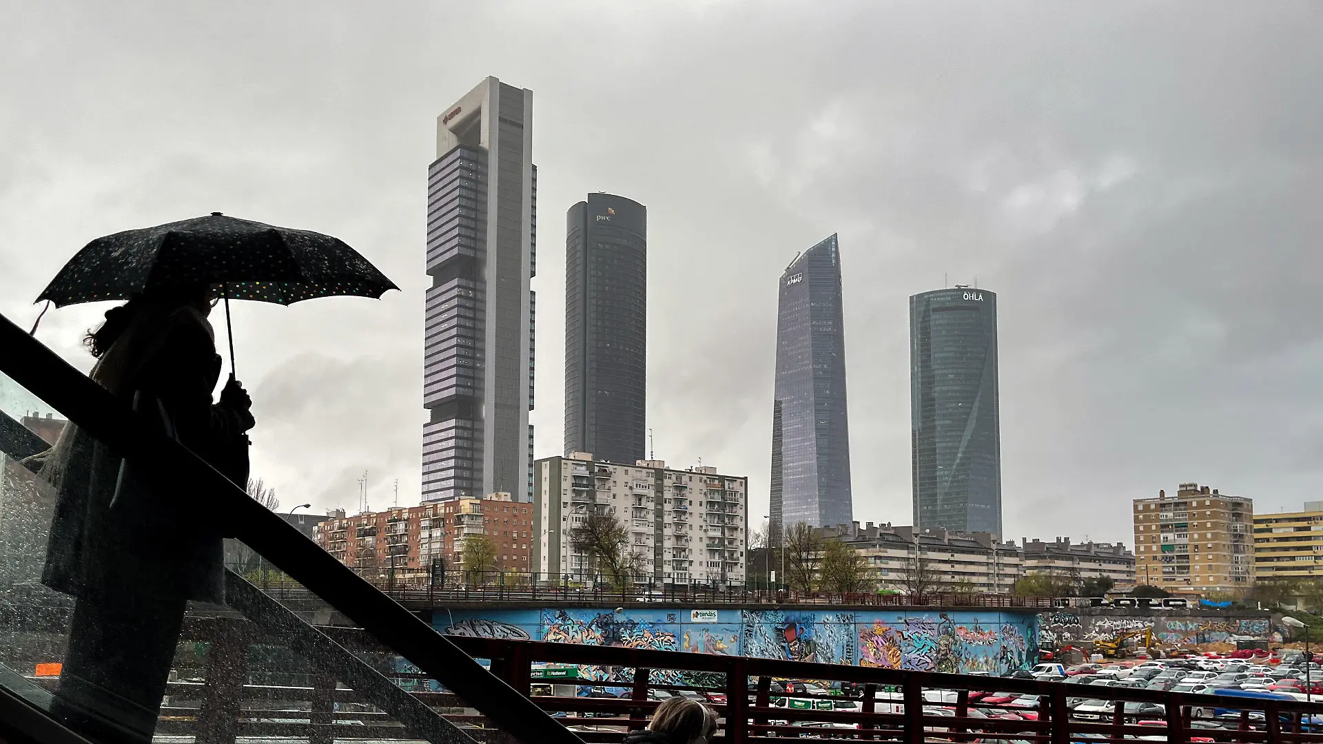 07.03.2024, Spanien, Madrid: Eine Frau mit Regenschirm fährt eine Rolltreppe vor der Kulisse der Cuatro Torres Business Area hinab. Die Meteorologen sagen auch für die nächsten Tage kühles und feuchtes Wetter für die Hauptstadt voraus. Foto: Jan Woitas/dpa +++ dpa-Bildfunk +++