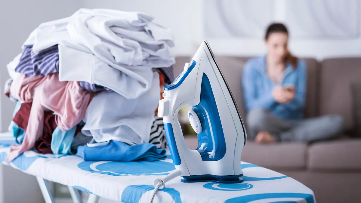 Lazy careless housewife on the couch at home and a pile of laundry on the ironing board on the foreground, boring household chores concept
