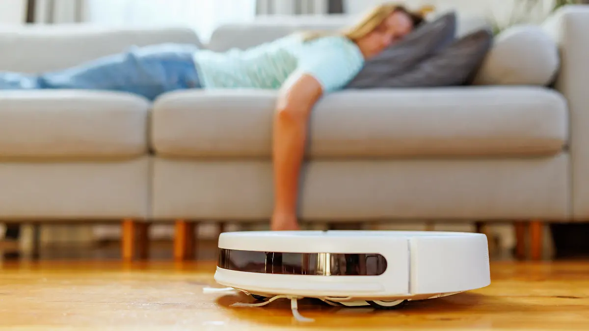 A woman takes a peaceful nap on the couch while the robotic vacuum cleaner quietly cleans the hardwood floor in a bright living room.