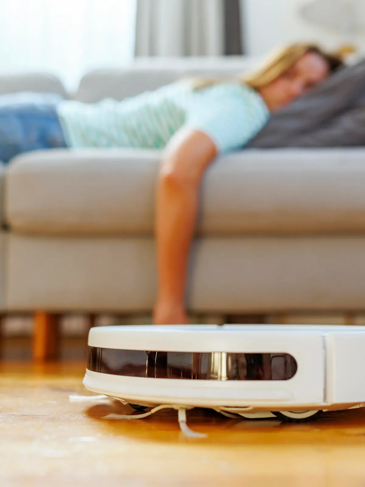 A woman takes a peaceful nap on the couch while the robotic vacuum cleaner quietly cleans the hardwood floor in a bright living room.
