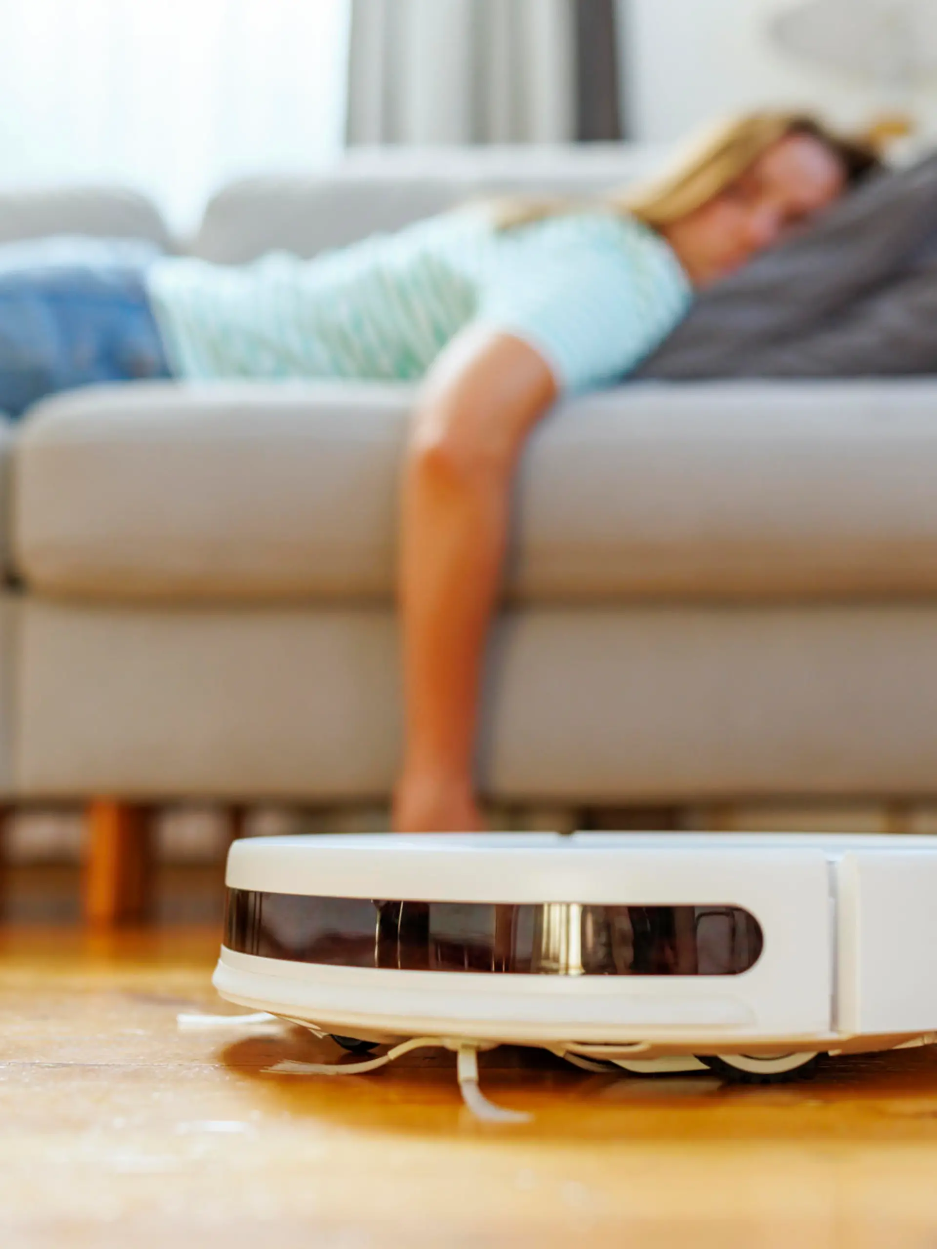 A woman takes a peaceful nap on the couch while the robotic vacuum cleaner quietly cleans the hardwood floor in a bright living room.