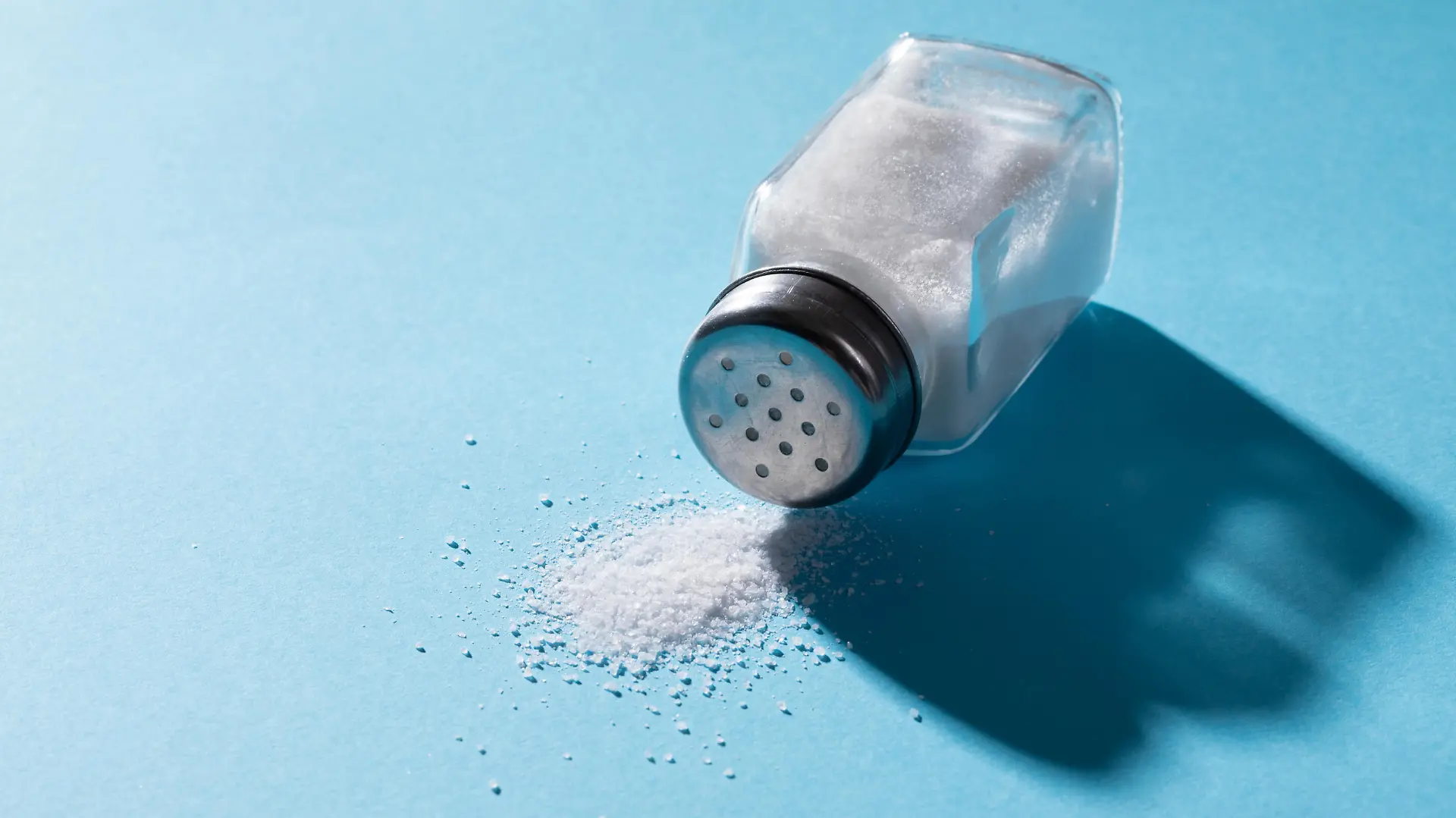 High angle close-up of salt spilling from salt shaker on blue background with copy space. unaltered, ingredient, food, shadow, rock salt and seasoning.