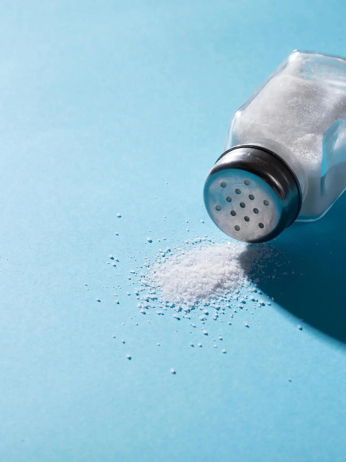 High angle close-up of salt spilling from salt shaker on blue background with copy space. unaltered, ingredient, food, shadow, rock salt and seasoning.