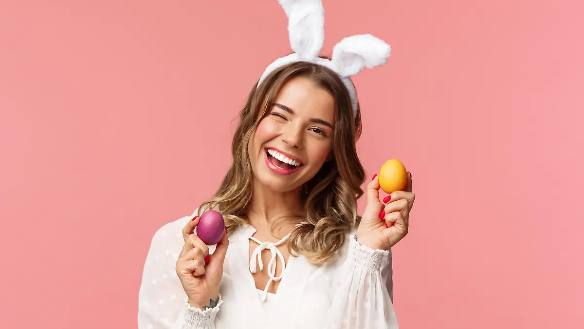 Holidays, spring and party concept. Cheerful good-looking blond woman celebrating Easter day in rabbit ears, holding two painted eggs and wink camera, smiling happily, pink background.