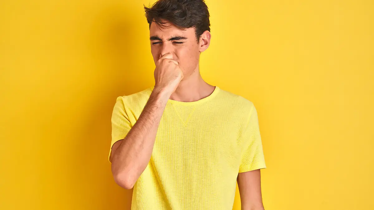 Teenager boy wearing yellow t-shirt over isolated background smelling something stinky and disgusting, intolerable smell, holding breath with fingers on nose. Bad smells concept.