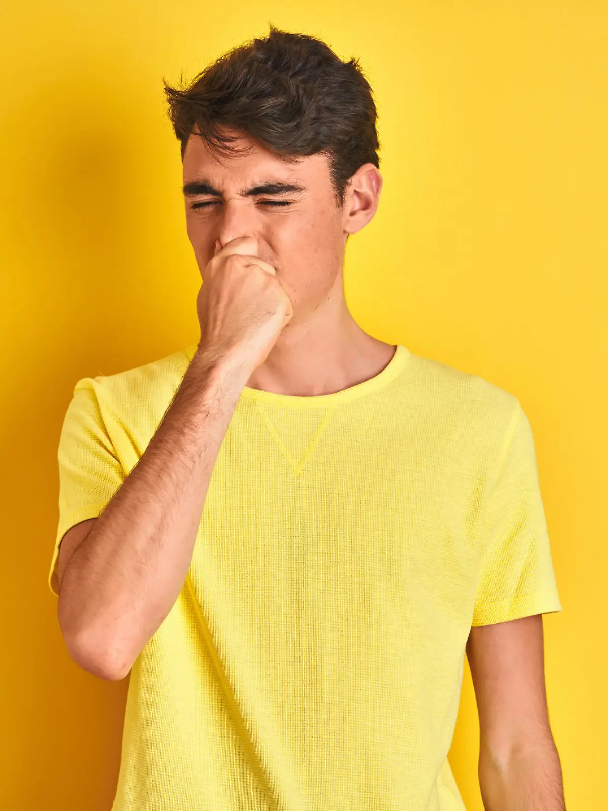 Teenager boy wearing yellow t-shirt over isolated background smelling something stinky and disgusting, intolerable smell, holding breath with fingers on nose. Bad smells concept.