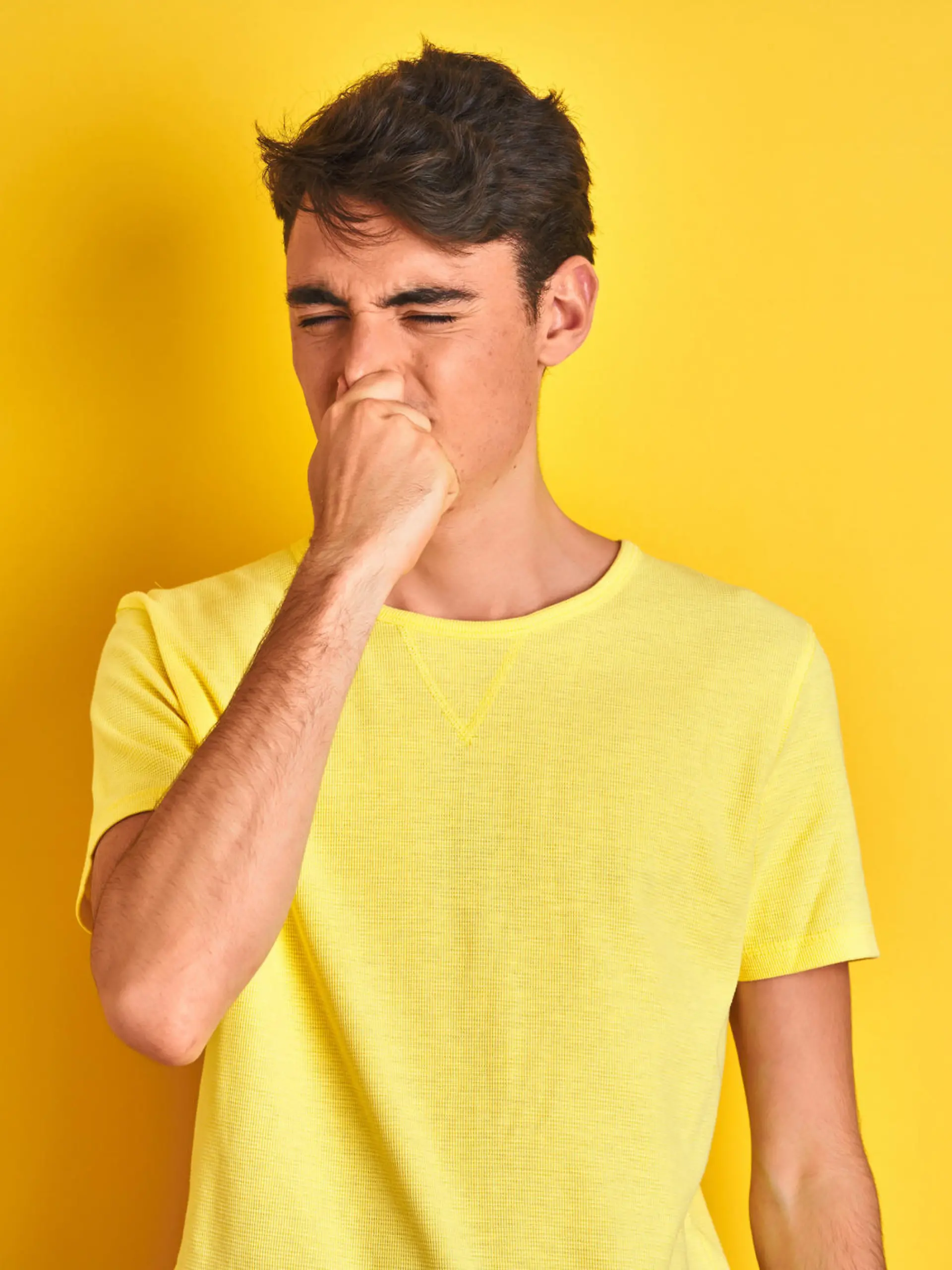 Teenager boy wearing yellow t-shirt over isolated background smelling something stinky and disgusting, intolerable smell, holding breath with fingers on nose. Bad smells concept.