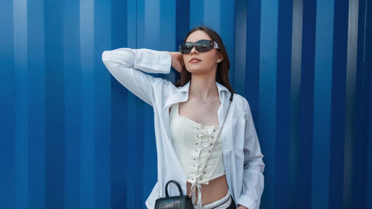 Fashionable beautiful young teenager girl with sunglasses in stylish clothes with a shirt, corset and handbag stands near a blue metal wall