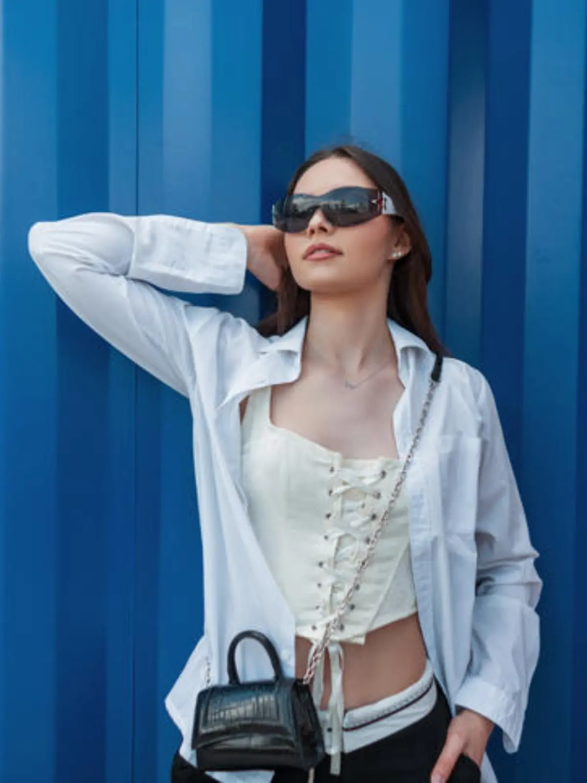 Fashionable beautiful young teenager girl with sunglasses in stylish clothes with a shirt, corset and handbag stands near a blue metal wall