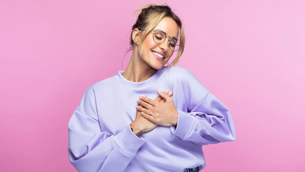Smiling woman with eyes closed keeping hands on heart while standing against pink background.