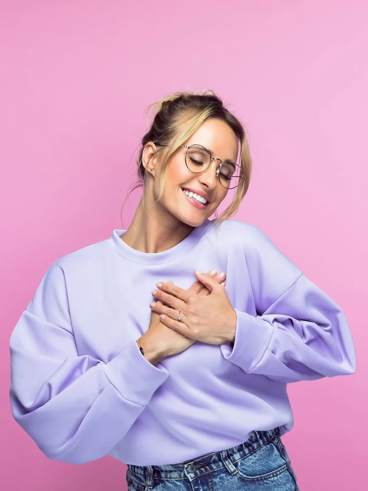 Smiling woman with eyes closed keeping hands on heart while standing against pink background.