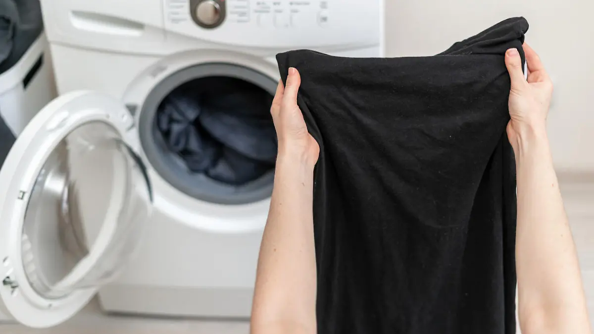 Cropped photo of laundry process. Woman holding black clothes in her hands against white washing machine inside light flat interior