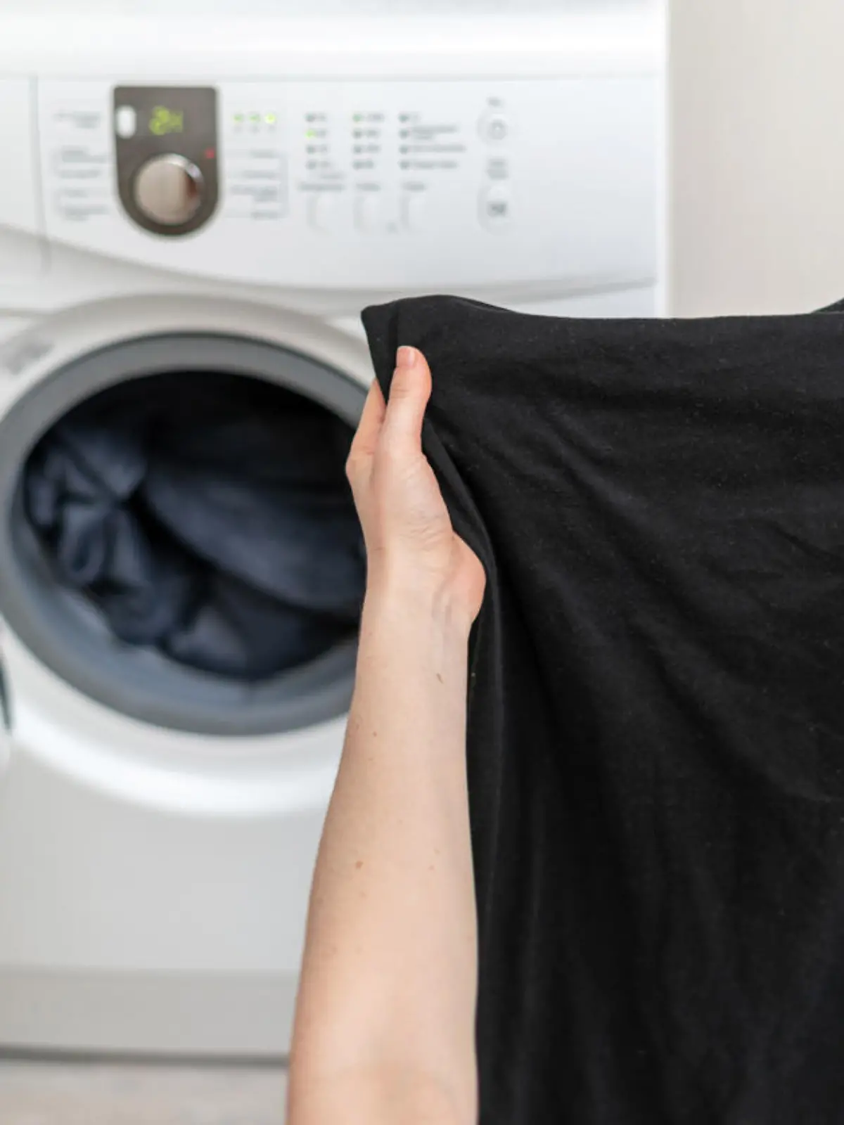 Cropped photo of laundry process. Woman holding black clothes in her hands against white washing machine inside light flat interior