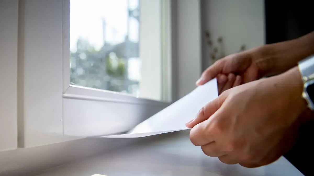 Frauenhände halten ein Stück Papier fest, welches im unteren Rahmen eines Fensters eingeklemmt wurde.