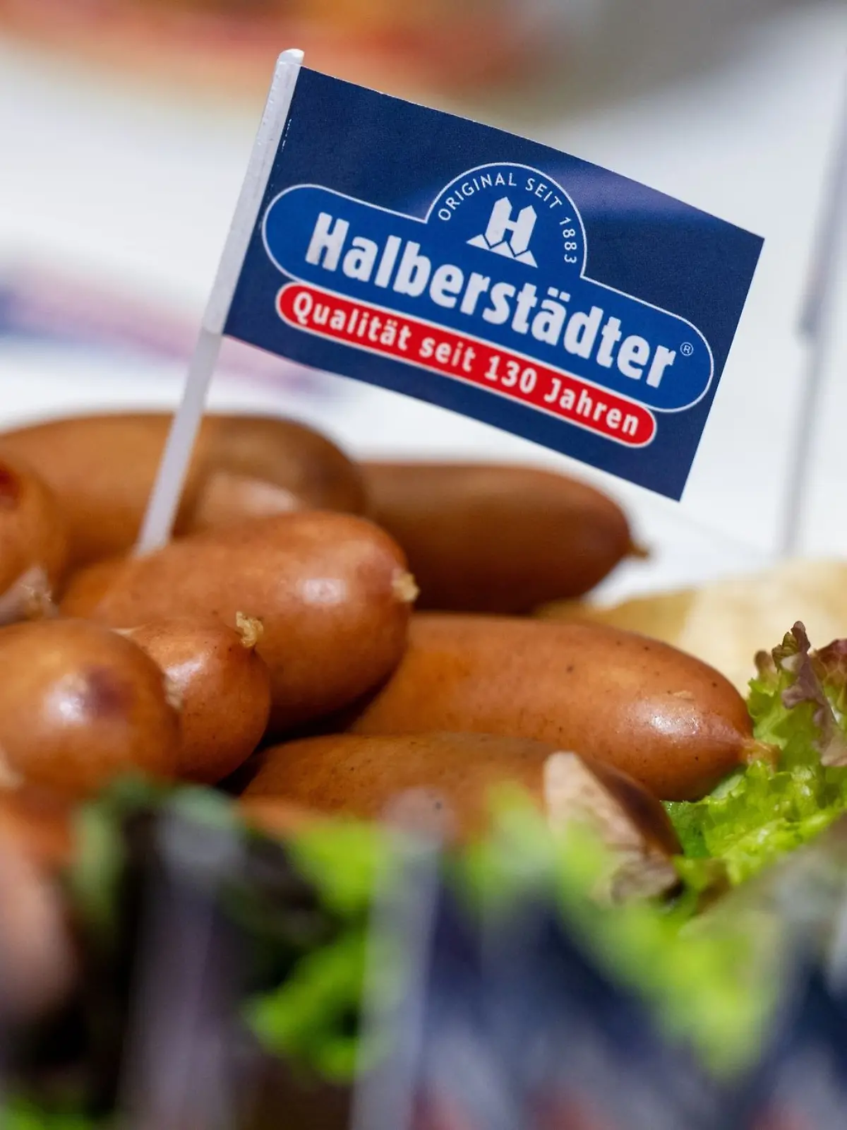 Ein Papierfähnchen mit dem Logo der Marke Halberstädter steckt in einem Würstchen am Stand des Herstellers auf der Grünen Woche.
