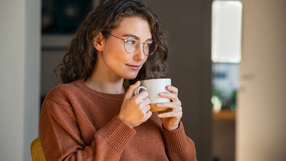 Pensive woman sitting in armchair and drinking hot coffee at home. Thoughtful young woman wearing eyeglasses drinking a cup of tea while thinking. Pretty contemplative girl with sweater relaxing at home while drinking purifying herbal tea during autumn, looking through the window.